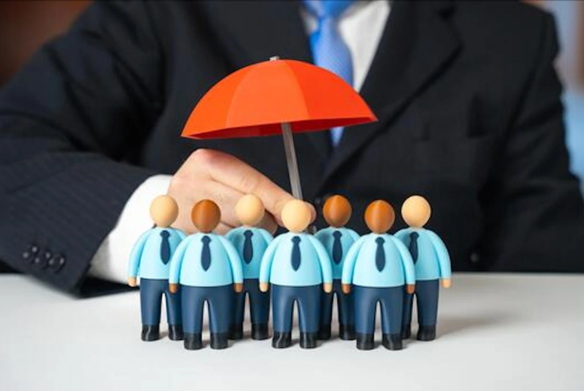 Figurines de collaborateurs en costume sur un oficina blanc, protégés par un parapluie rouge tenu par una personne en costume profesional — métaphore de la cobertura multirriesgo empresa